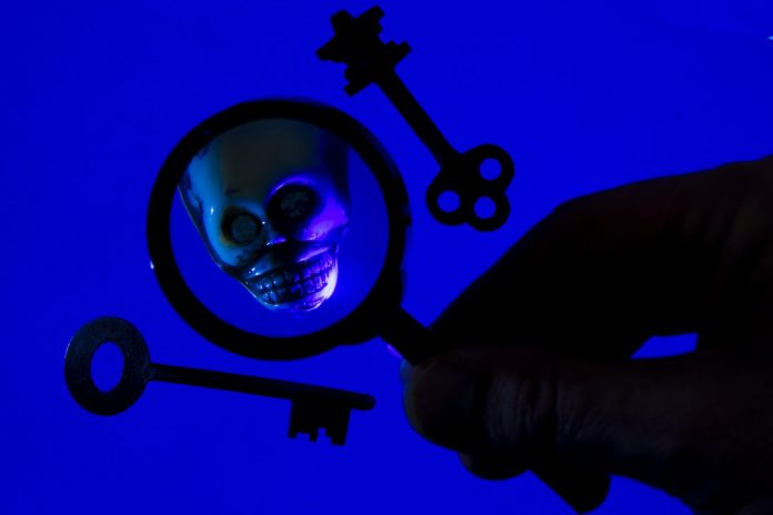 On a blue background, two old fashioned keys are displayed on either side of a shadowy hand holding magnifying glass, which reveals a magnified skull.